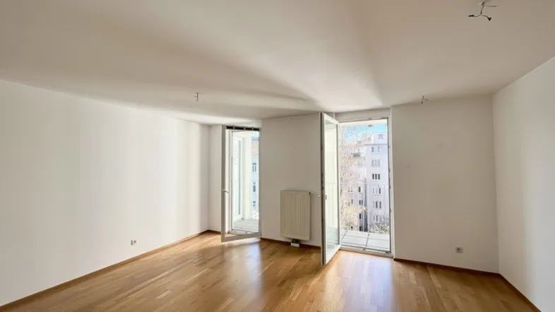 Helles, leeres Zimmer mit Parkettboden und Zugang zu zwei Balkonen, ideal für Wohn- oder Schlafbereich.