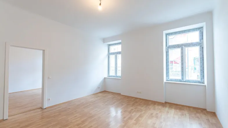 Helles, leeres Zimmer mit Parkettboden und zwei großen Fenstern, bereit zur individuellen Gestaltung.