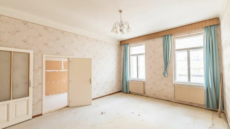 Geräumiger Wohnraum mit floraler Tapete, zwei Fenstern und Kronleuchter, bereit zur Renovierung.