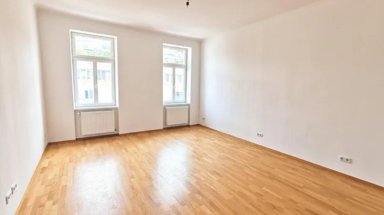 Helles, leeres Zimmer mit Parkettboden und zwei großen Fenstern, bereit zur individuellen Gestaltung.