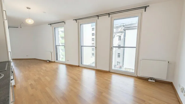 Helles, leeres Zimmer mit Parkettboden und großen Fenstern, die viel Tageslicht hereinlassen.