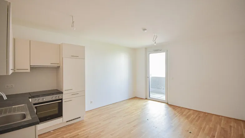 Heller offener Wohnbereich mit integrierter Küchenzeile, Parkettboden und Zugang zu einem Balkon.