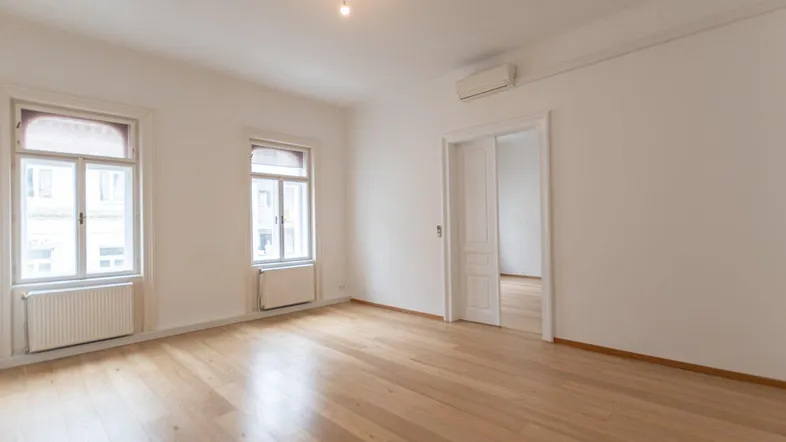 Helles Zimmer mit Holzboden und zwei Fenstern, ideal als Wohn- oder Schlafbereich.
