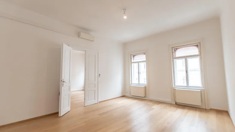 Helles, leeres Zimmer mit Parkettboden und zwei Fenstern, bereit zur individuellen Gestaltung.
