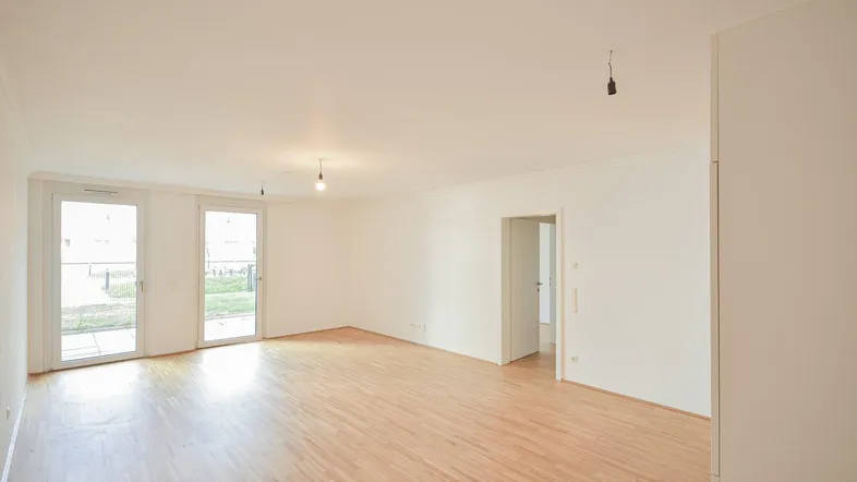 Helles, leeres Wohnzimmer mit Parkettboden und Zugang zu einem Außenbereich durch große Fenstertüren.