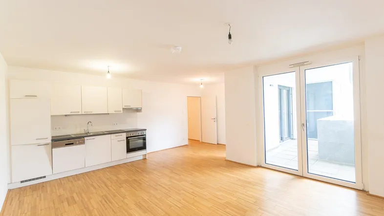 Heller offener Wohnbereich mit integrierter Küche, Parkettboden und Zugang zum Balkon.