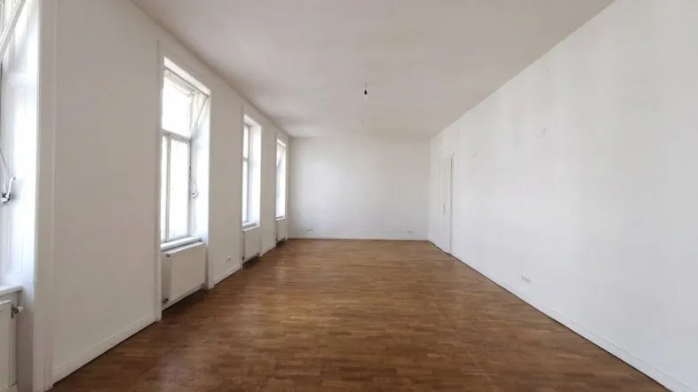Langer, heller Raum mit mehreren Fenstern, die den Holzboden und die weißen Wände beleuchten.