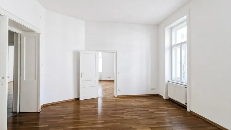 Heller, leerer Raum mit weiß gestrichenen Wänden, Holzboden und großen Fenstern, die viel Tageslicht hereinlassen.