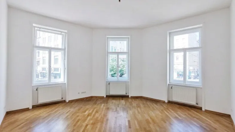 Heller, leerer Raum mit Holzboden und drei großen Fenstern, die viel Tageslicht hereinlassen.