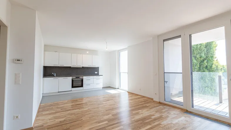 Heller offener Wohnbereich mit Parkettboden, integrierter Küche und Zugang zu einem Balkon.