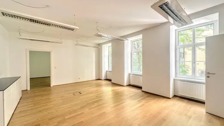Geräumiger Büroraum mit hellem Holzfußboden, hohen Decken und großen Fenstern, die viel Tageslicht hereinlassen.