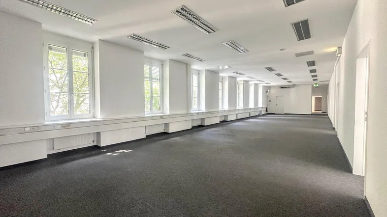 Großes, leeres Büro mit Teppichboden, vielen Fenstern und Deckenbeleuchtung, ideal für flexible Arbeitsplatzgestaltung.
