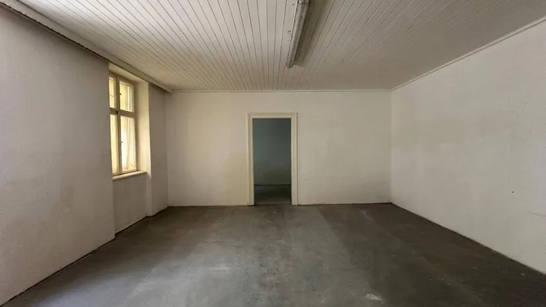 Leerer Raum mit Betonboden, weiß gestrichenen Wänden und einem Fenster, das Tageslicht hereinlässt.