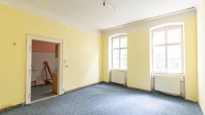 Geräumiger Raum mit gelben Wänden, blauem Teppichboden und zwei Fenstern, bereit zur Renovierung.
