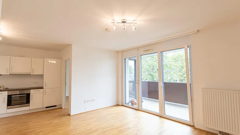 Heller offener Wohnbereich mit integrierter Küche, Parkettboden und Zugang zum Balkon.