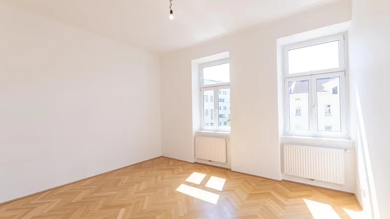 Helles Zimmer mit Fischgrätparkett, weißen Wänden und zwei großen Fenstern, durch die Sonnenlicht fällt.