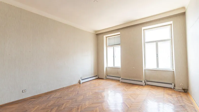 Geräumiges Zimmer mit Fischgrätparkett und zwei großen Fenstern, das eine Modernisierung benötigt.