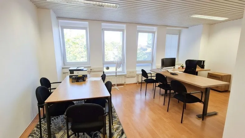 Helles Büro mit mehreren Arbeitsplätzen, Holzboden und großen Fenstern mit Blick ins Grüne.