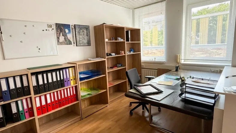 Heller Büroraum mit Schreibtisch, Bürostuhl, Holzregalen und Fenstern, die viel Tageslicht hereinlassen.