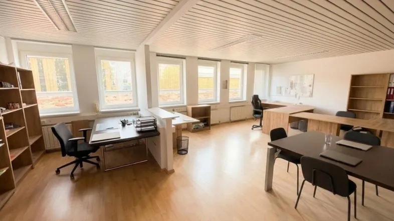 Geräumiges Büro mit mehreren Arbeitsplätzen, großen Fenstern und hellem Laminatboden, ideal für produktives Arbeiten.
