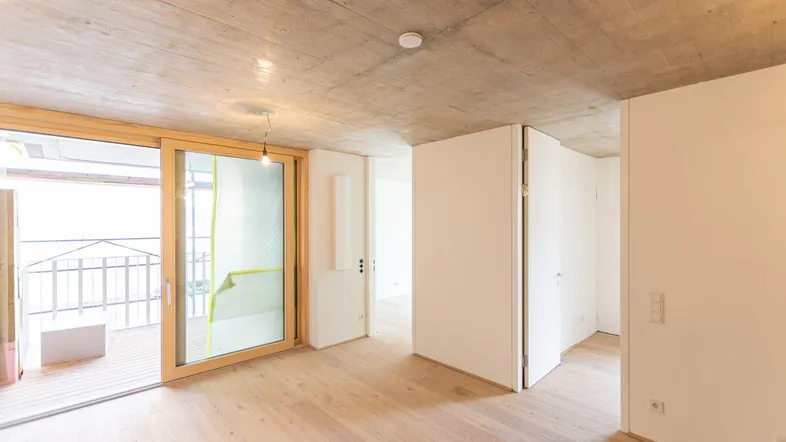 Lichtdurchfluteter Raum mit Schiebetür zum Balkon, Holzboden und moderner Betonoptikdecke.