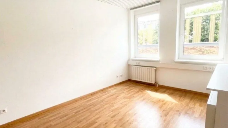 Heller Büroraum mit zwei großen Fenstern, Holzfußboden und weiß gestrichenen Wänden.