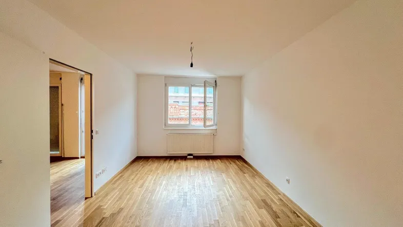 Helles, leeres Zimmer mit Parkettboden und einem Fenster, das Tageslicht hereinlässt.