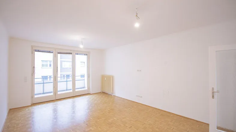 Helles, leeres Wohnzimmer mit Parkettboden und Zugang zu einem Balkon.