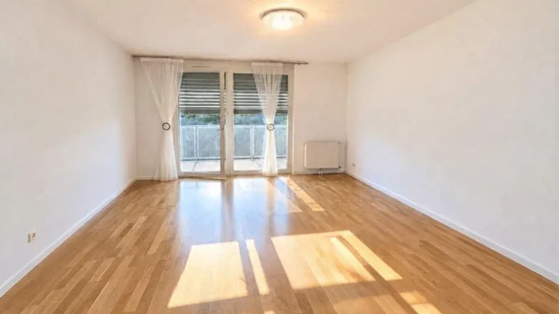 Helles, leeres Wohnzimmer mit Holzboden und Zugang zu einem Balkon, durchflutet von Sonnenlicht.