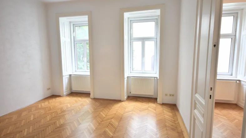 Helles Zimmer mit Fischgrätparkett und zwei großen Fenstern, ideal als Wohn- oder Schlafzimmer.