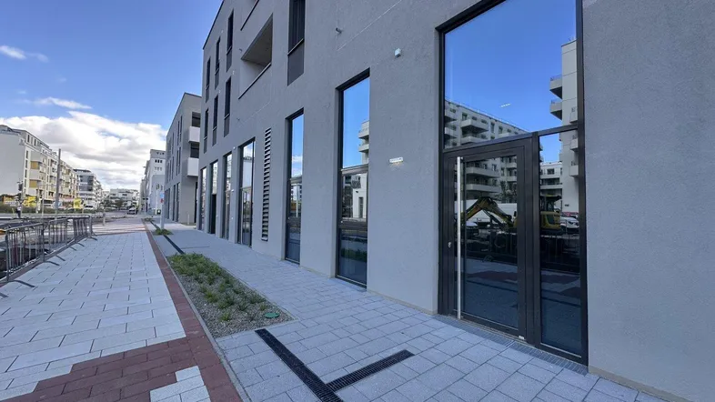 Moderne Gebäudefassade mit großen, reflektierenden Fenstern und einem Eingangsbereich an einem Gehweg.