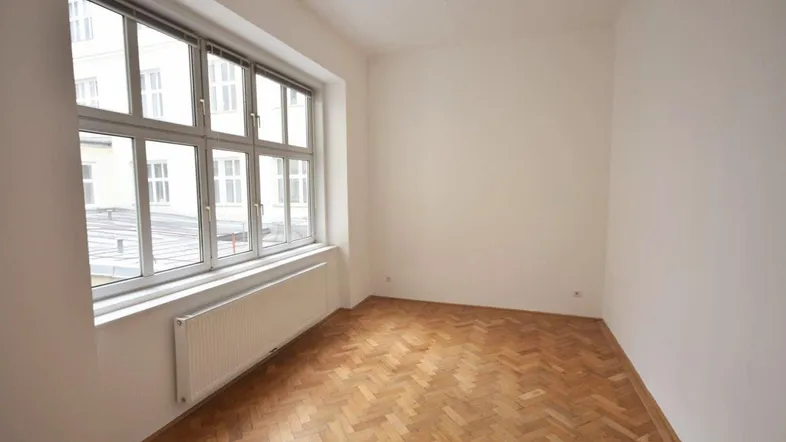 Helles Zimmer mit Fischgrätparkett, weißen Wänden und großem Fenster, ideal als Wohn- oder Schlafzimmer.