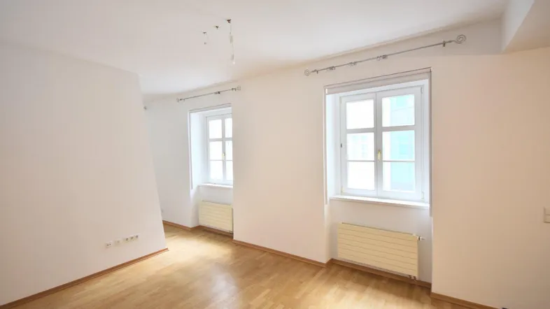 Helles, leeres Zimmer mit Parkettboden und zwei Fenstern, bereit zur individuellen Gestaltung.