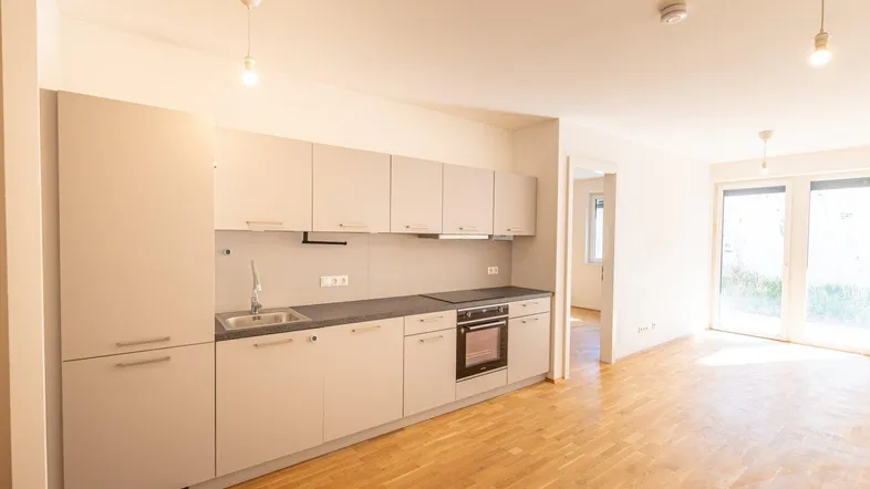 Moderne Einbauküche mit hellgrauen Schränken, dunkler Arbeitsplatte und Holzboden in offenem Wohnbereich.