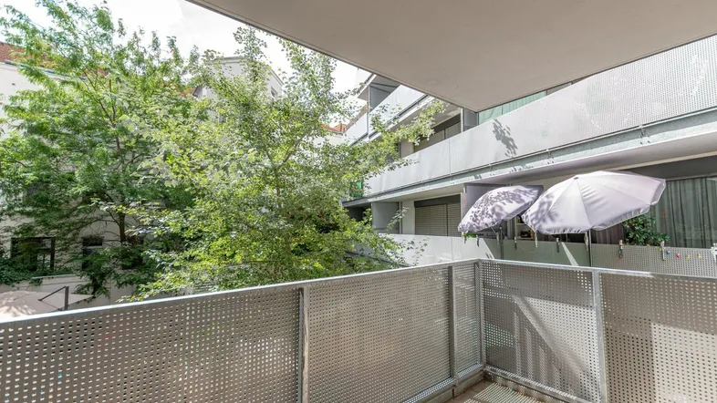 Balkon mit Metallgeländer und Blick auf grüne Bäume und umliegende Gebäude.