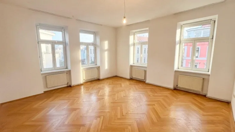 Helles Zimmer mit Fischgrätparkett, weißen Wänden und mehreren Fenstern, ideal als Wohnzimmer.