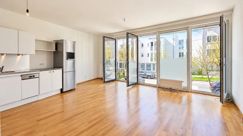 Heller offener Wohnbereich mit integrierter Küche, Parkettboden und großen Fenstern zur Straße.