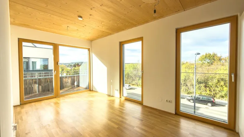 Helles Zimmer mit Holzboden und Holzdecke, großen Fenstern und Zugang zum Balkon.