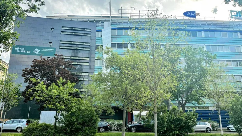 Moderne Bürogebäude-Fassade mit blauen Akzenten und großen Fenstern, umgeben von üppigem Grün.