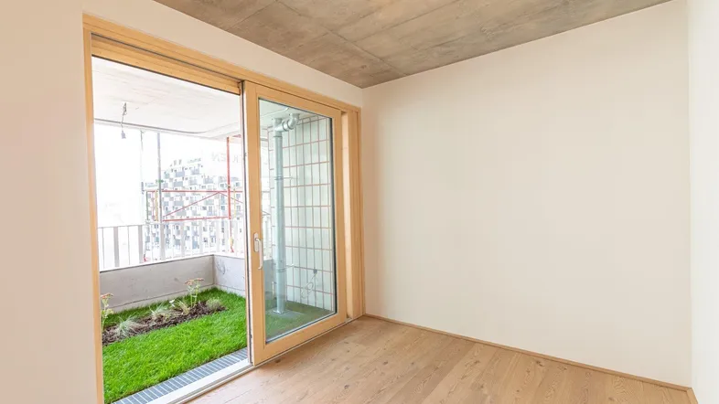 Heller Raum mit Holzboden, Sichtbetondecke und Zugang zu einem kleinen begrünten Außenbereich.