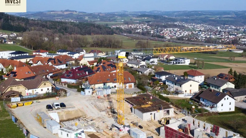Luftaufnahme einer Baustelle mit Kran, umgeben von Wohnhäusern und grüner Landschaft, mit einer Stadt in der Ferne.