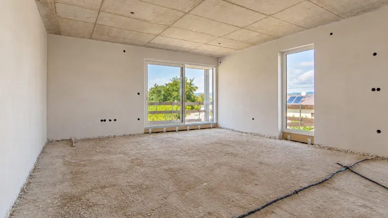 Unfertiger Raum in einem Neubau mit großen Fenstern, die viel Tageslicht hereinlassen und einen Blick ins Grüne bieten.