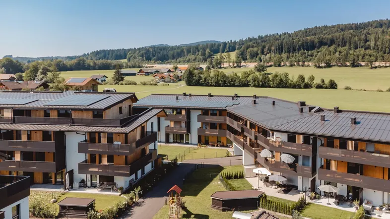 Moderne Wohnanlage mit mehreren Gebäuden, Balkonen, Grünflächen und ländlicher Umgebung unter blauem Himmel.