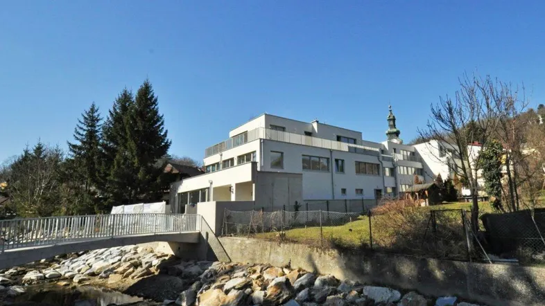 Modernes Gebäude mit Terrassen und Balkonen, gelegen an einem Bach mit Brücke und Blick auf eine Kirche.