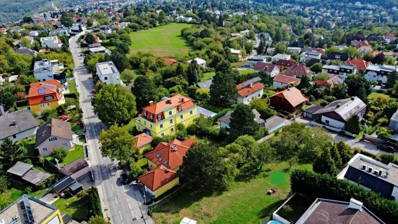 Luftaufnahme eines Wohngebiets mit grünen Hügeln und einem Baugrundstück in der Mitte.