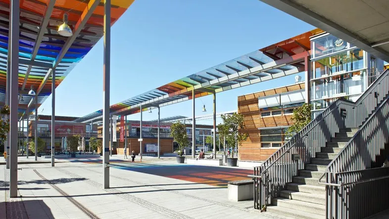 Weitläufiger Außenbereich eines modernen Gebäudekomplexes mit bunten Überdachungen, Treppen und einem Aufzug.