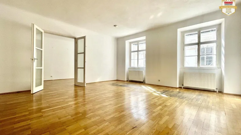 Geräumiges Zimmer mit hellem Parkettboden, weißen Wänden und hohen Fenstern, die viel Tageslicht hereinlassen.