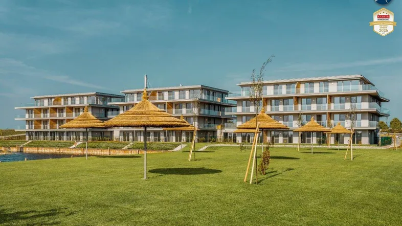 Moderne Wohngebäude am Seeufer mit weitläufiger Rasenfläche und Strohschirmen für entspannte Stunden.