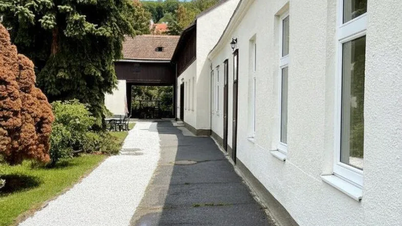 Weißes Gebäude mit langem Weg, umgeben von Bäumen und Grünflächen unter blauem Himmel.