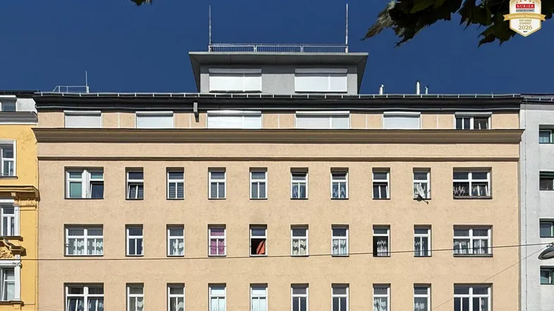 Fassade eines Mehrfamilienhauses mit zahlreichen Fenstern und einem modernen Dachaufbau unter blauem Himmel.